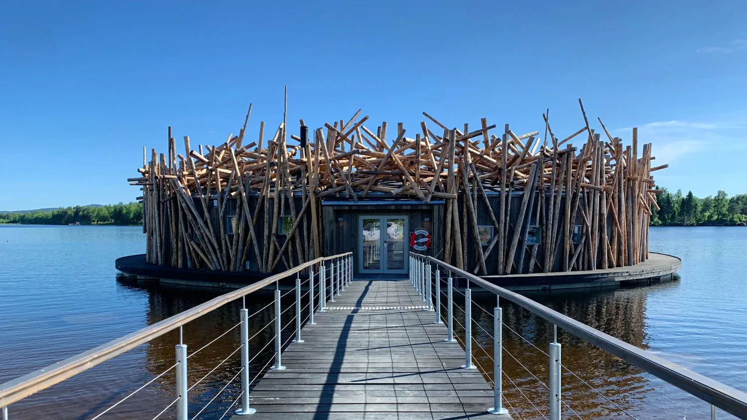 Circular Arctic Bath spa building with crisscross log exterior connected by a wooden walkway on a calm summer river