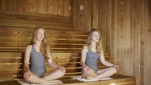 Two women in blue geometric swimsuits sitting in meditation pose inside a wooden sauna