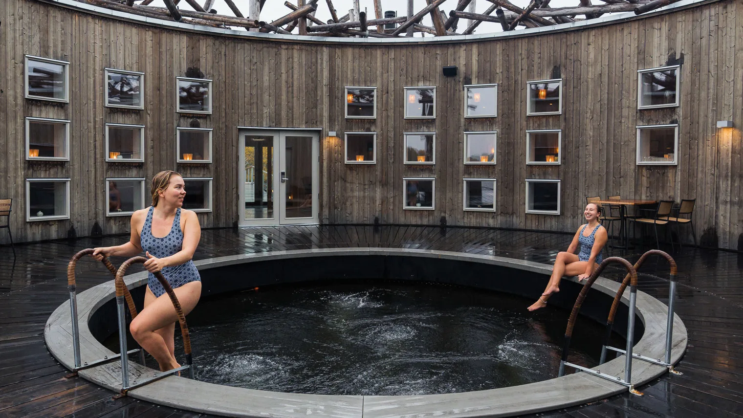 Two women in blue swimsuits at a circular outdoor cold plunge pool in a wooden log-walled courtyard