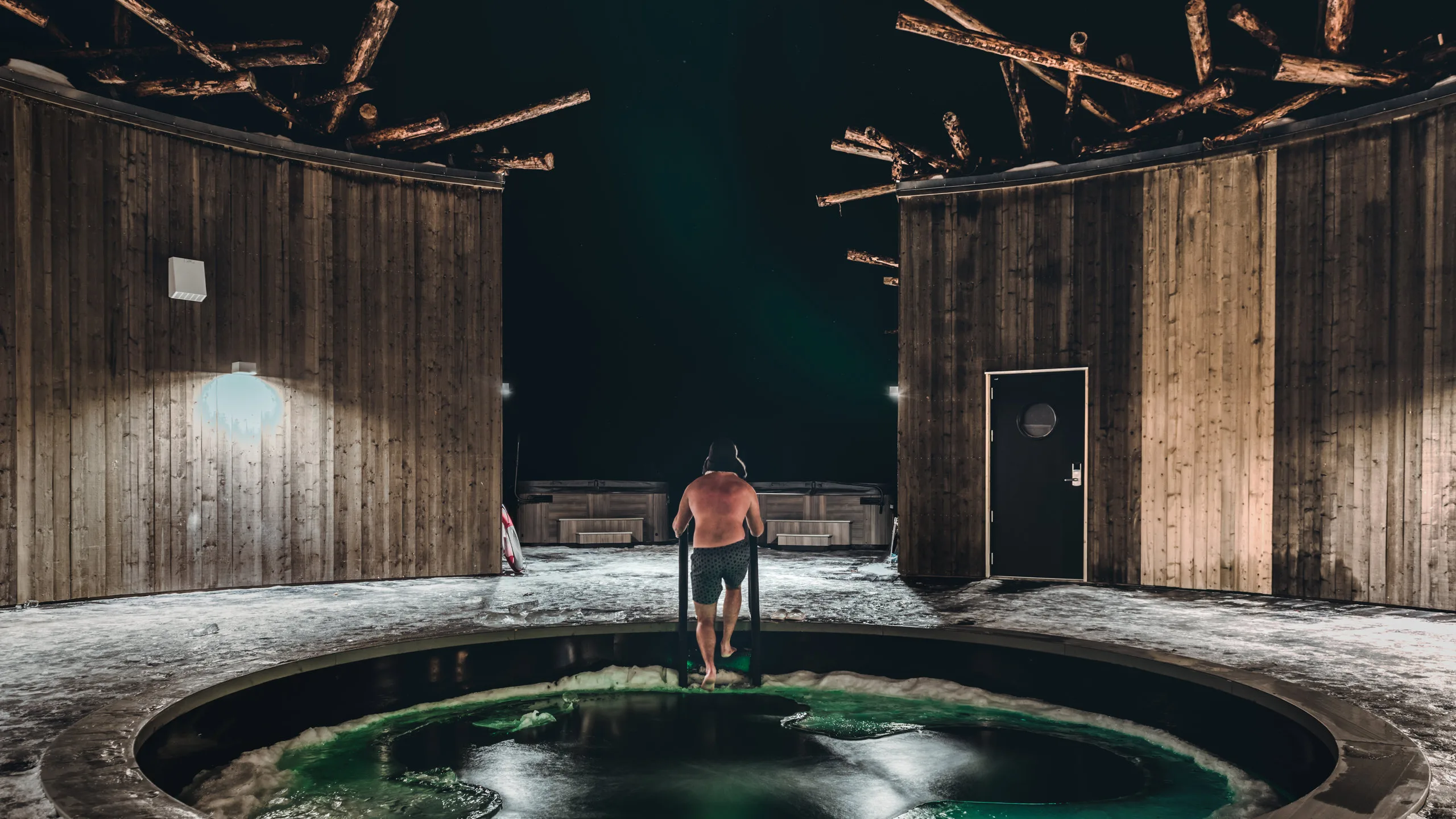 Man descending into the illuminated cold plunge at Arctic Bath's snowy circular courtyard at night
