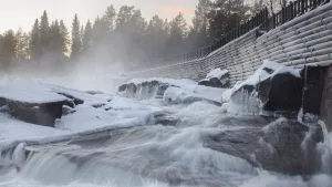 Long-exposure winter river rapids with snow-covered rocks and a curved log wall shrouded in mist