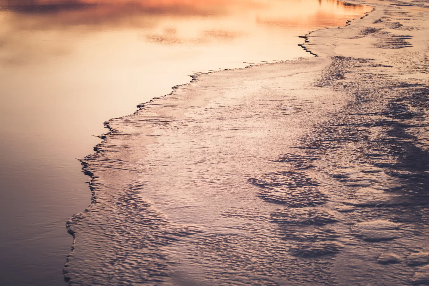 Close-up of frozen ice textures beside open water reflecting warm sunset pink hues
