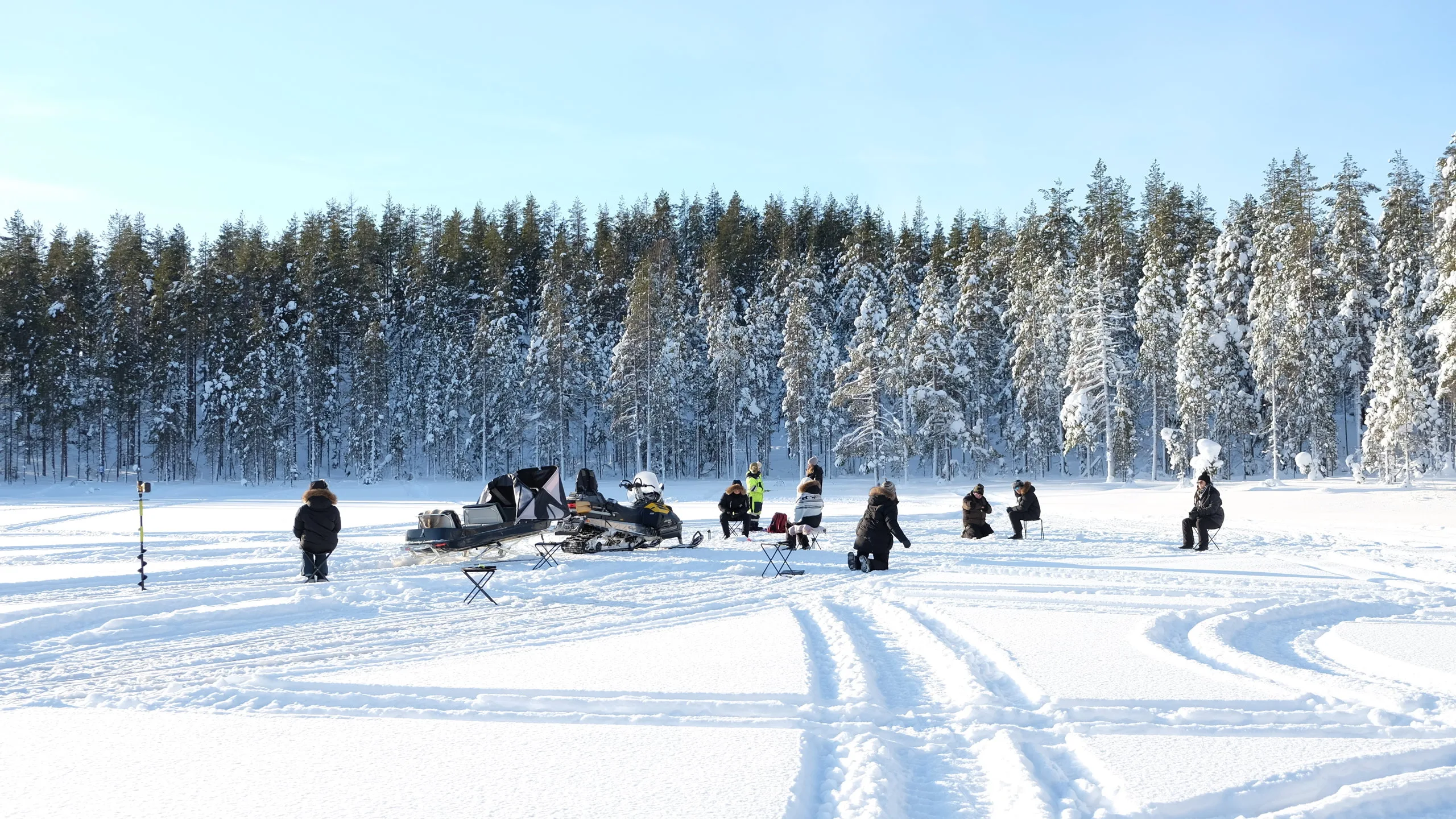 Group ice fishing on a snow-covered frozen lake with snowmobile tracks and forest backdrop