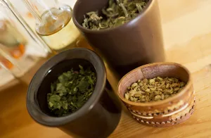 Dried herbs in ceramic jars and a carved wooden bowl with seeds on a light wooden table