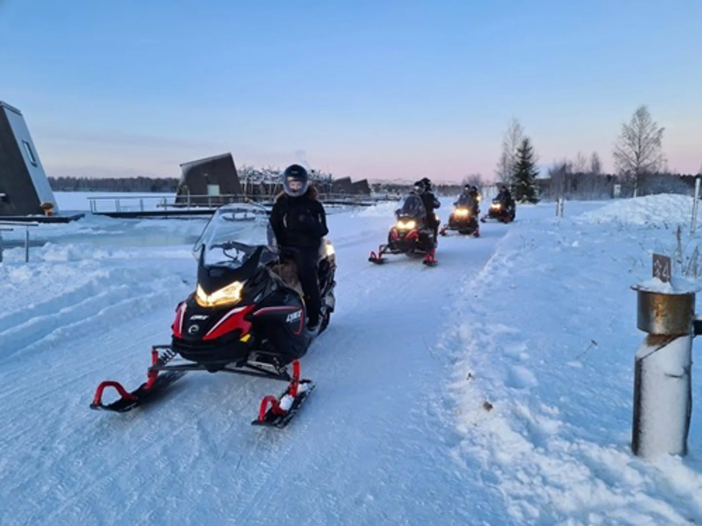 Line of snowmobiles departing from Arctic Bath past land cabins across a snowy landscape at twilight
