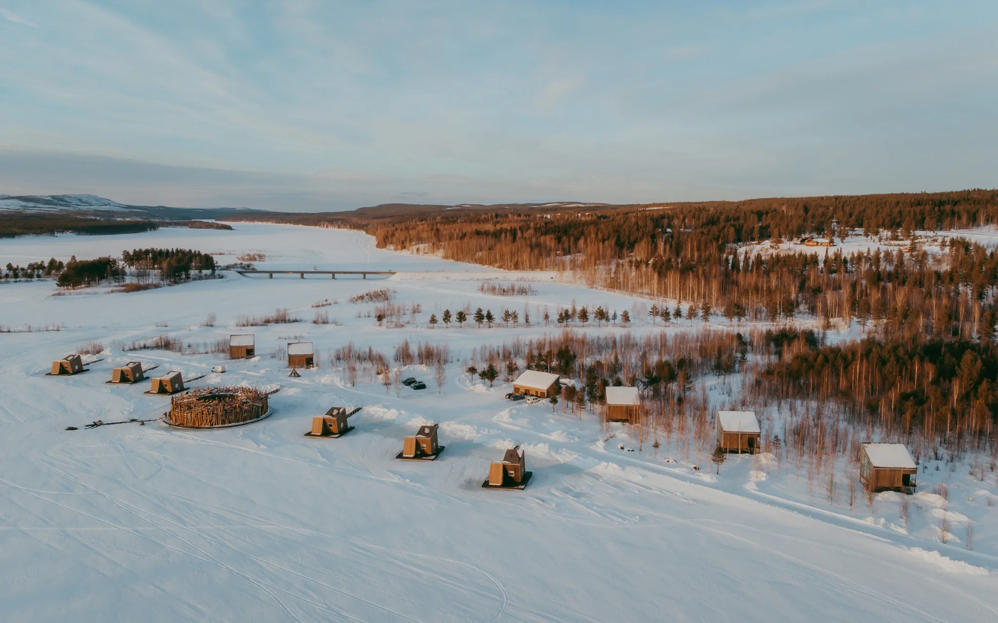 Aerial view of Arctic Bath resort at sunset with the circular main building, land cabins, and frozen Lule River