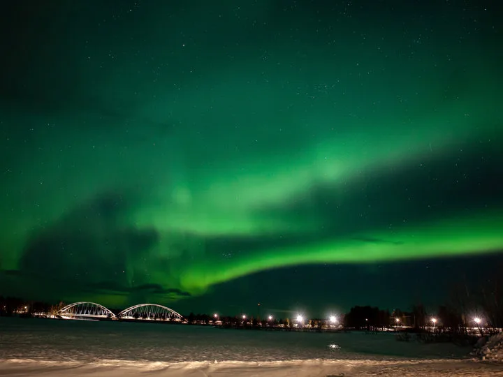 Green northern lights arching over a snow-covered riverbank with an illuminated bridge and distant lights