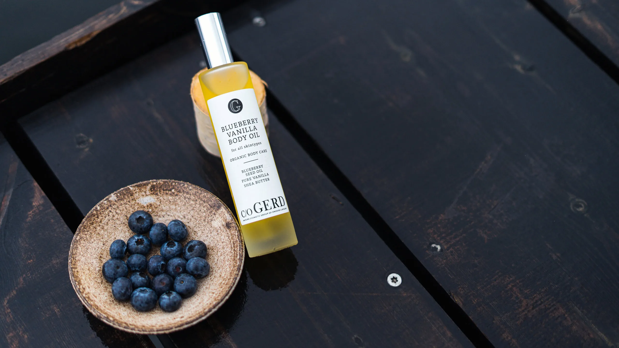 C/O Gerd blueberry body oil bottle beside a small bowl of fresh blueberries on dark wood
