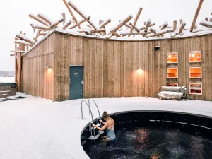 Guest entering the circular ice bath at Arctic Bath, with snow-covered wooden walls and a log-topped roof glowing warmly behind