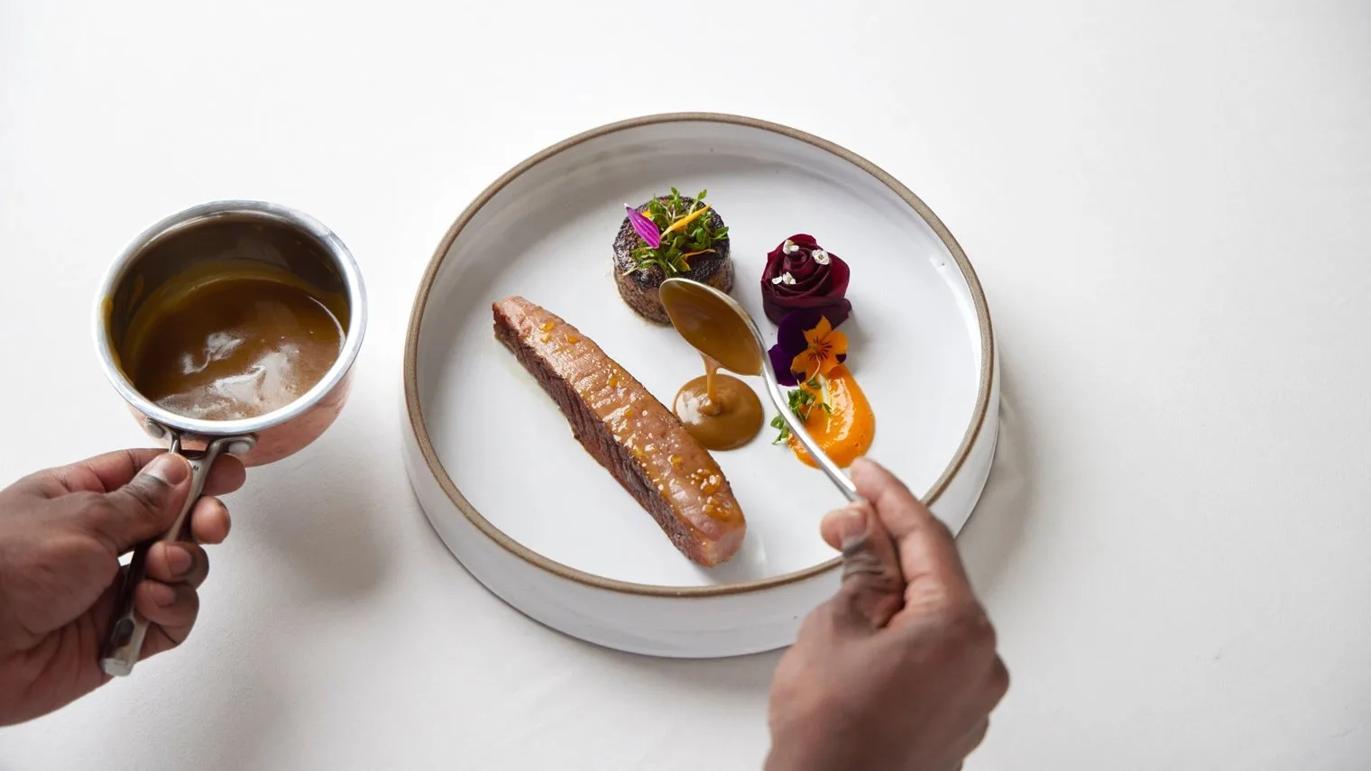 Chef drizzling brown sauce onto a fine dining plate with seared meat and garnishes
