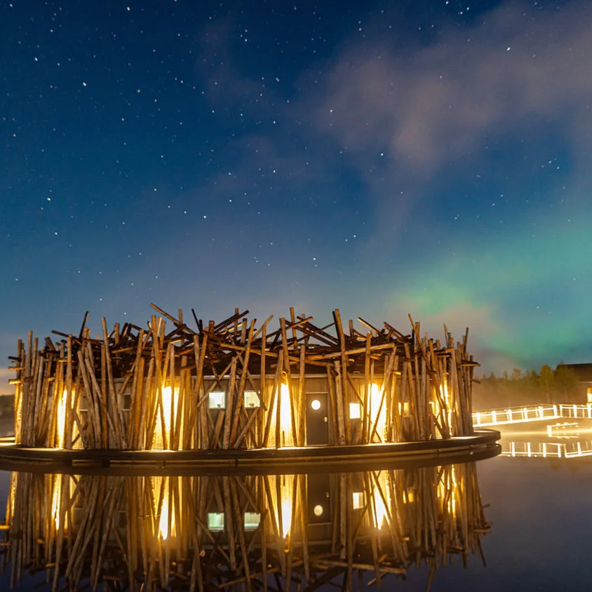 Circular Arctic Bath spa building at night with warm interior light and faint aurora reflected on the water