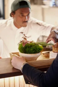 Chef arranging moss, pastry and greenery on a wooden tray beside a guest's hand on a dark counter